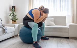 Frustrated overweight woman sitting on exercise ball in living room with her head in her hands