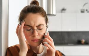Young woman visibly frustrated with her hand at her temple while on hold with her doctors office